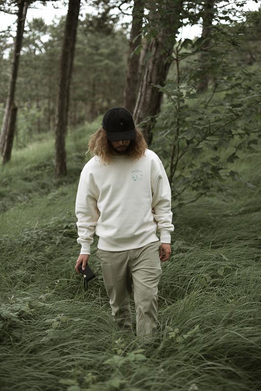 Worry Way Less Crewneck Man Standing In The Green Woods With Trees And Grass On The Background Wearing A Crewneck Of Organic Cotton With Small Screenprinted Design Chest Front